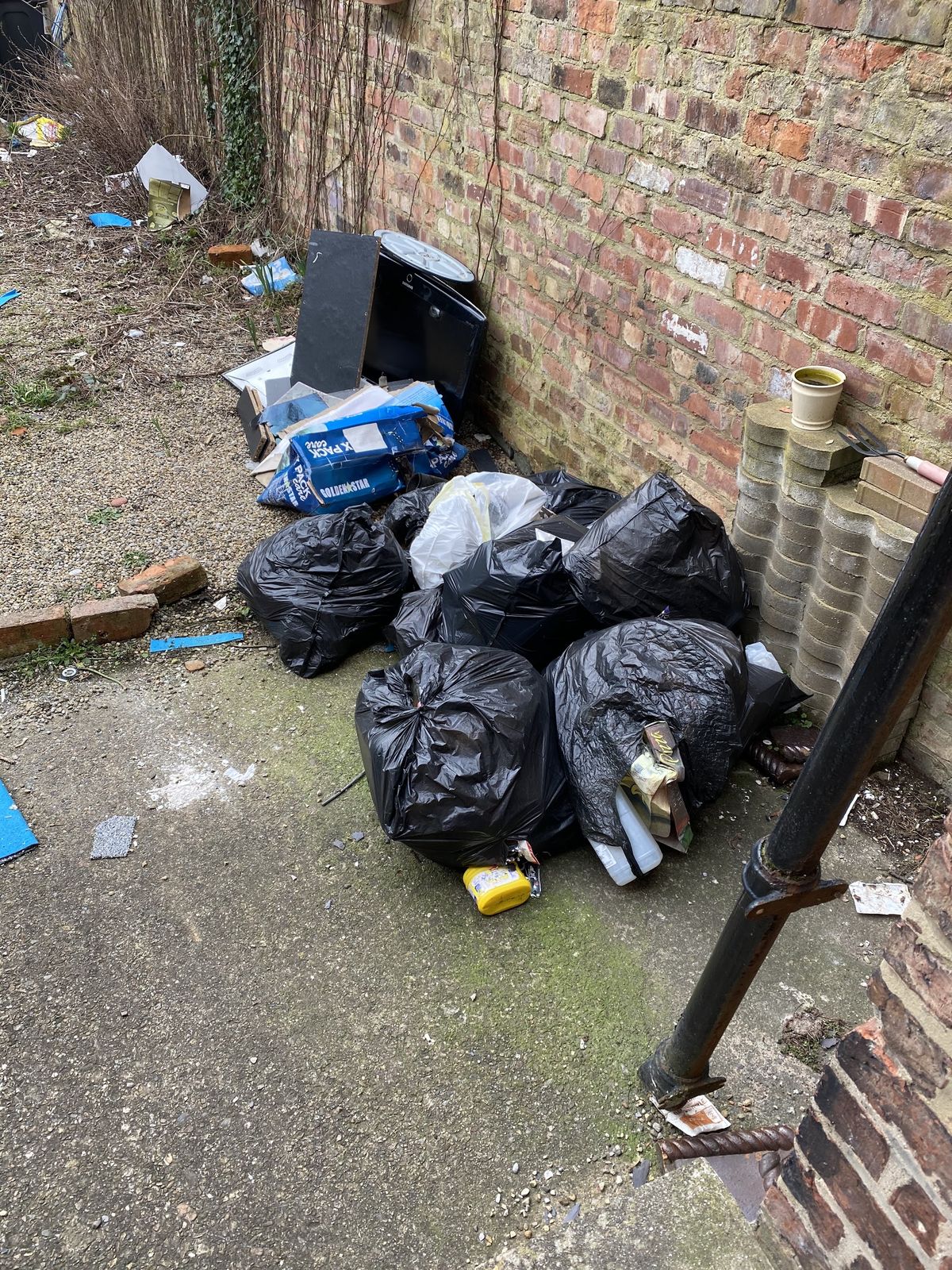 Rubbish bags and debris piled against a brick wall in a back yard