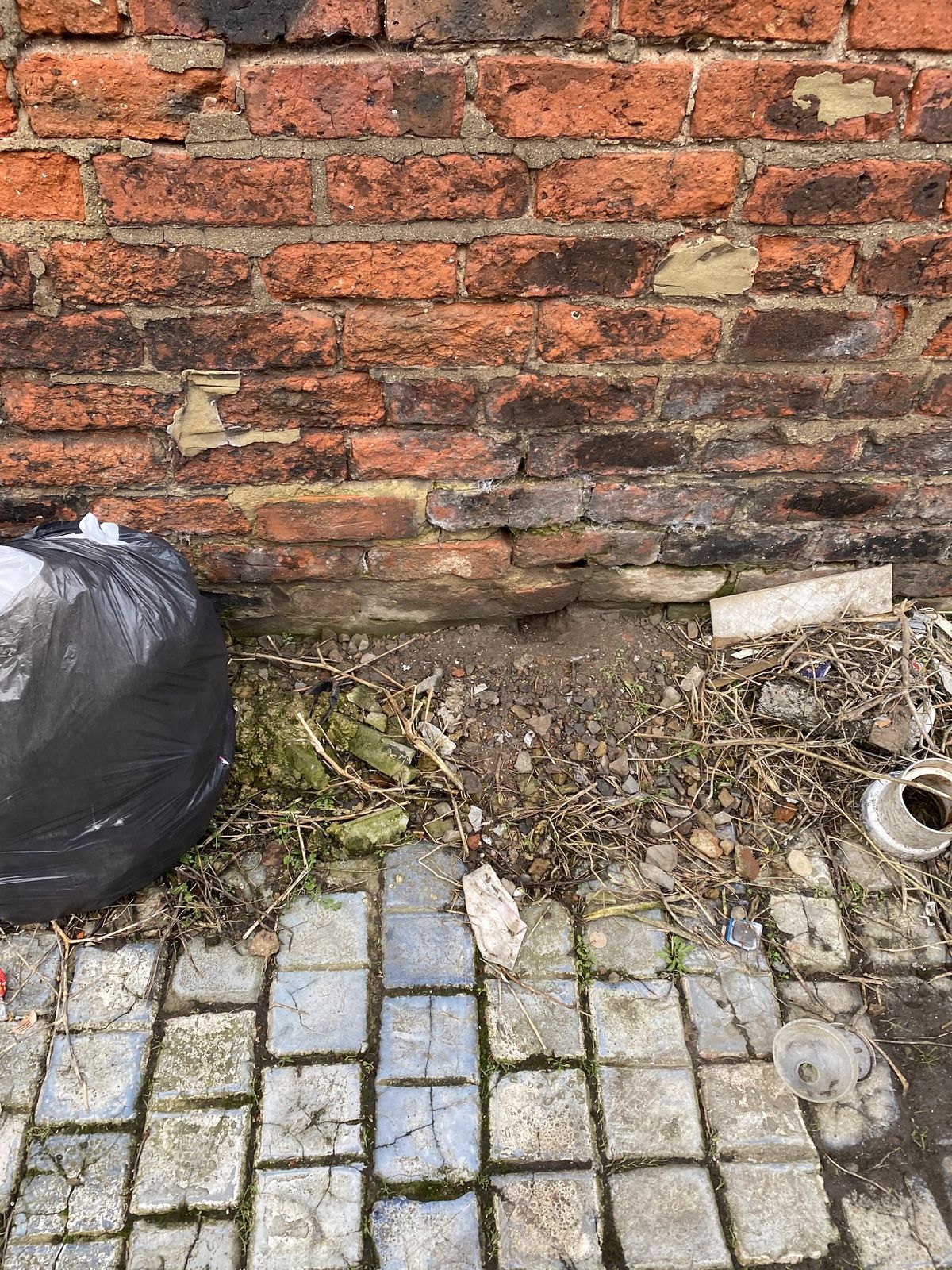 The base of a brick wall with a bin bag and debris on block paving